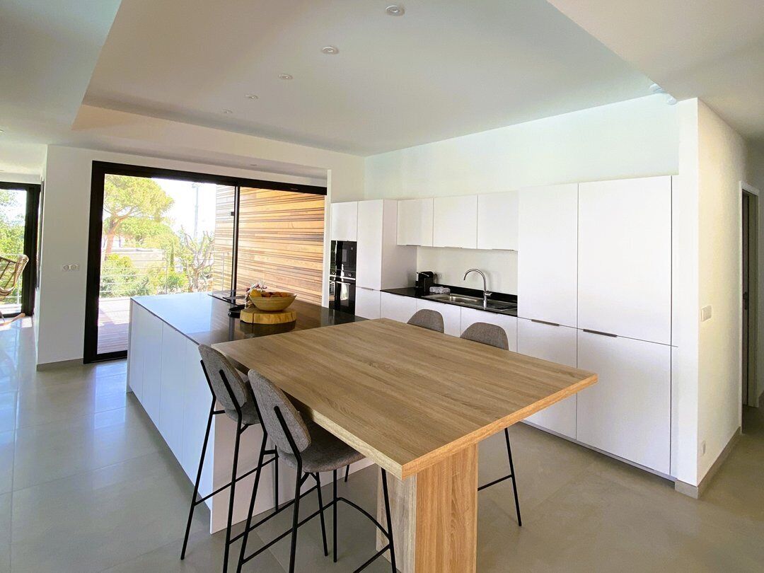 Une cuisine moderne avec des meubles blancs, une table à manger en bois et une grande fenêtre donnant sur un jardin.