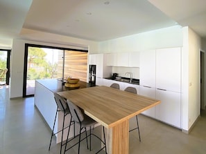 A modern kitchen with white cabinets, a wooden dining table, and a large window overlooking a garden. - Villa Maria 2. 12 pers, piscine chauffée, 5 min plage (sainte lucie de porto vecchio)