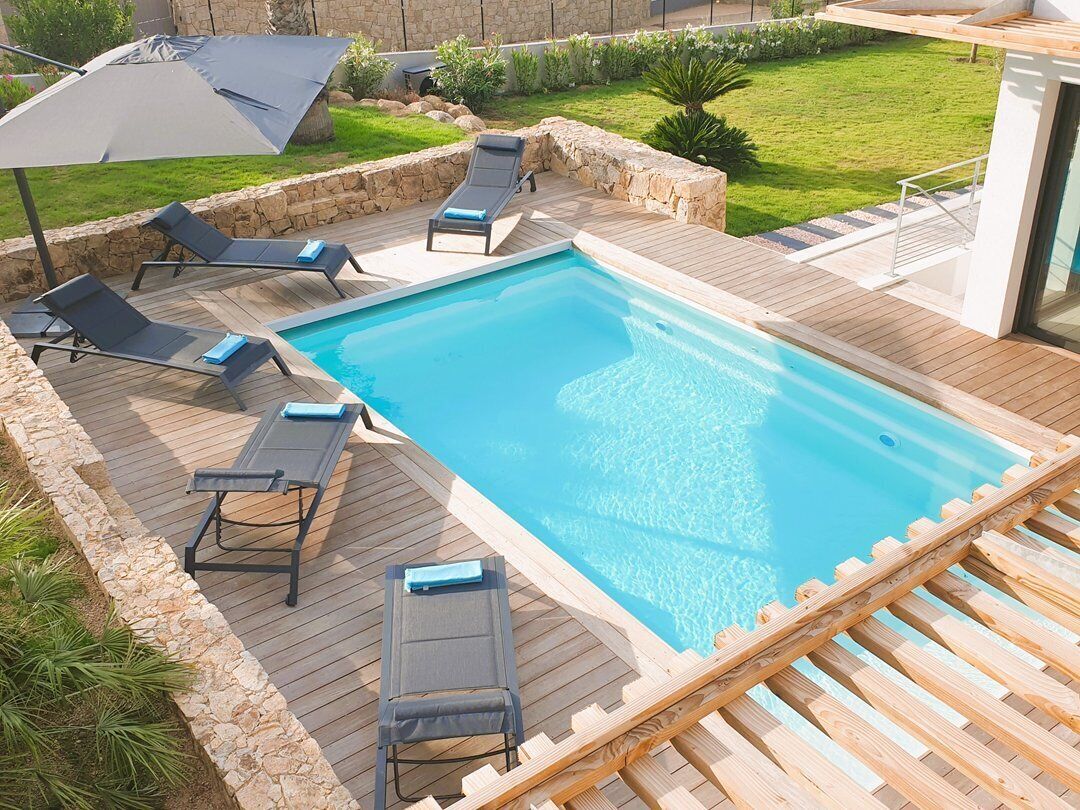 A rectangular swimming pool with a wooden deck, surrounded by lounge chairs and a stone wall.