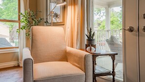 Une pièce avec un fauteuil beige, une petite table en bois sur laquelle est posée une plante, un lampadaire et une vue sur une véranda dotée d’une balustrade.