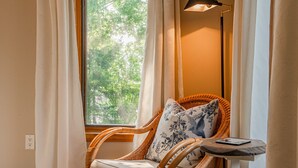 Une chaise en osier, agrémentée d’un coussin et d’un oreiller à motifs floraux, placée près d’une fenêtre ornée de rideaux blancs.