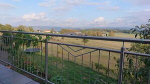 Un balcon doté d’une rambarde en métal, donnant sur un champ verdoyant, un trampoline et offrant une vue lointaine sur des maisons et des collines.