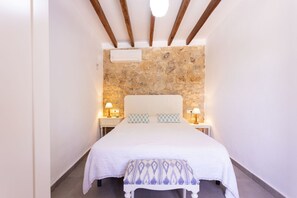 A narrow hotel room with a stone wall, a white bed, two bedside lamps, and a small bench.