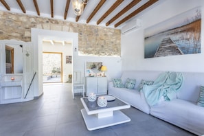 A modern living room with a stone wall, a white sofa, a coffee table with decorative items, and a painting of a dock. - Village house with pool, 3 bedrooms, 2 bathrooms, barbecue (Campanet)