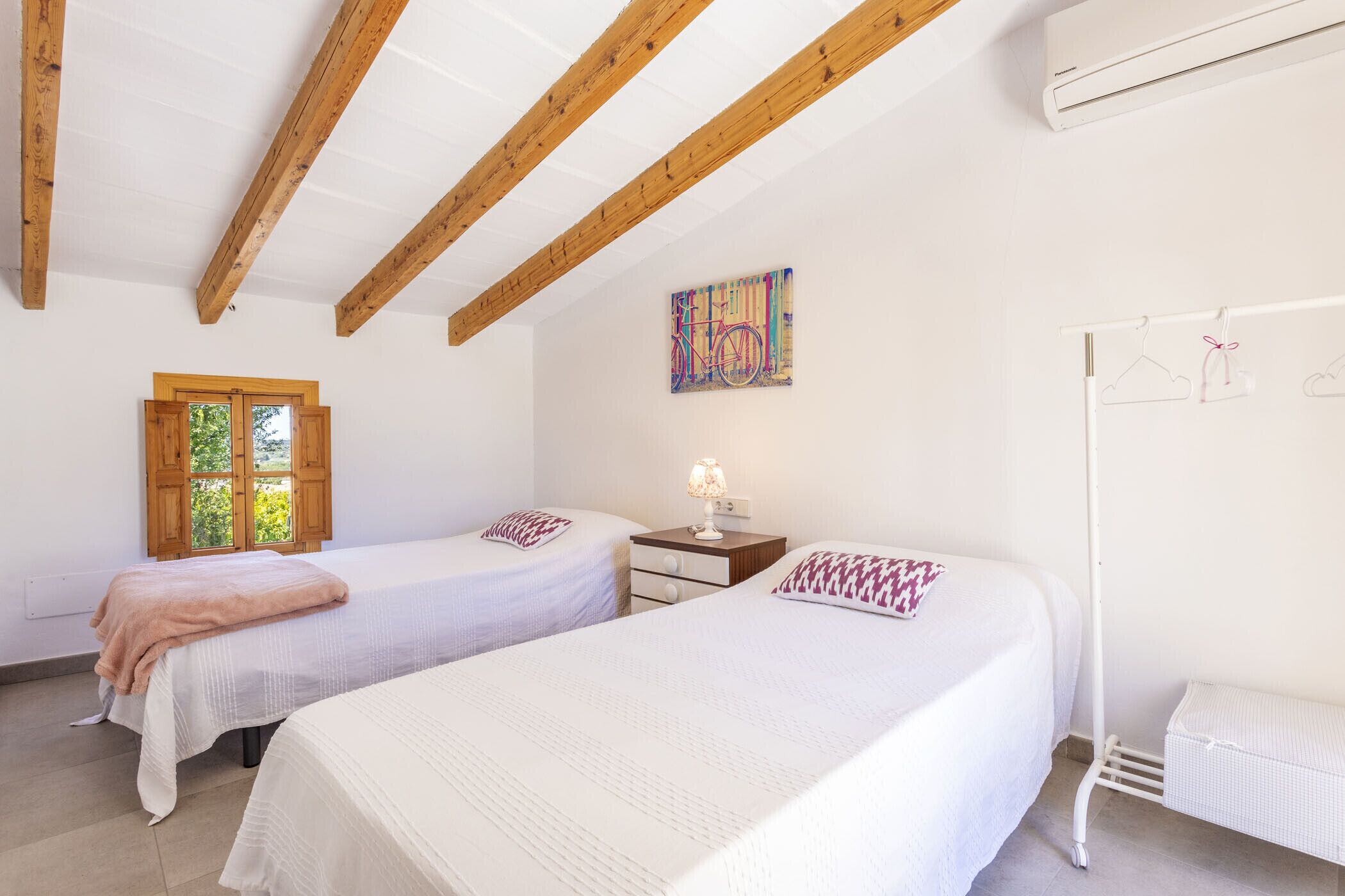 A bedroom with two beds, wooden beams on the ceiling, a window with shutters, a wall-mounted air conditioner, and a hanging clothes rack.