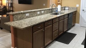 A kitchen with granite countertops, brown cabinets, a stainless steel sink, and a dishwasher. There is a flat-screen TV mounted on the wall and two pendant lights above the counter. - Bay View, Downtown Traverse City Condo (Traverse City)