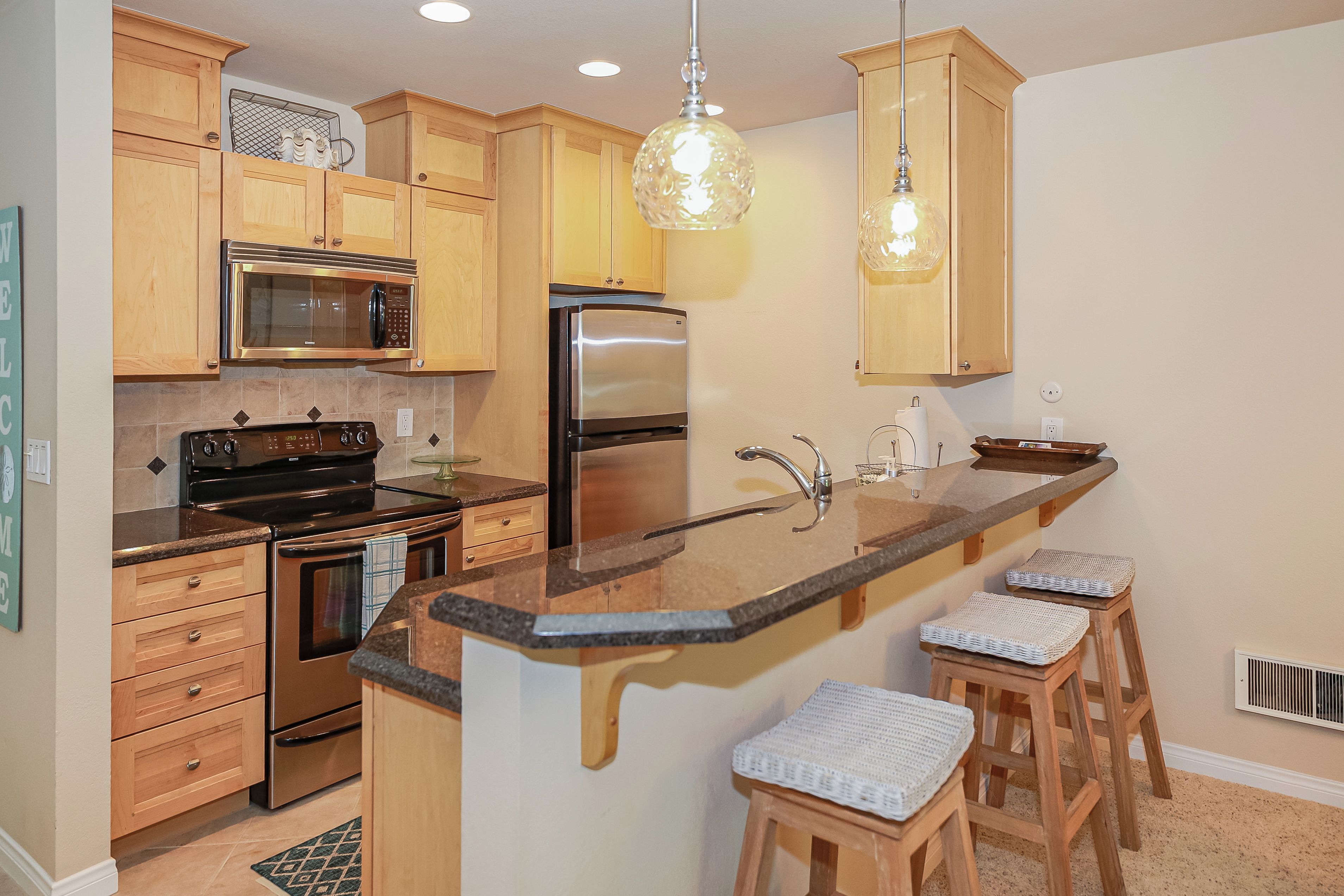 A kitchen with wooden cabinets, a stainless steel refrigerator, a microwave, an oven, a sink, and bar stools.
