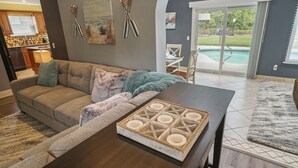 A modern living room with a dark wooden coffee table, a beige sofa with decorative pillows, and a view of a backyard with a pool. - HEATED Saltwater POOL, Harry Potter, BBQ, Lake, Fishing, Gazebo, Games & Firepit (St. Augustine)
