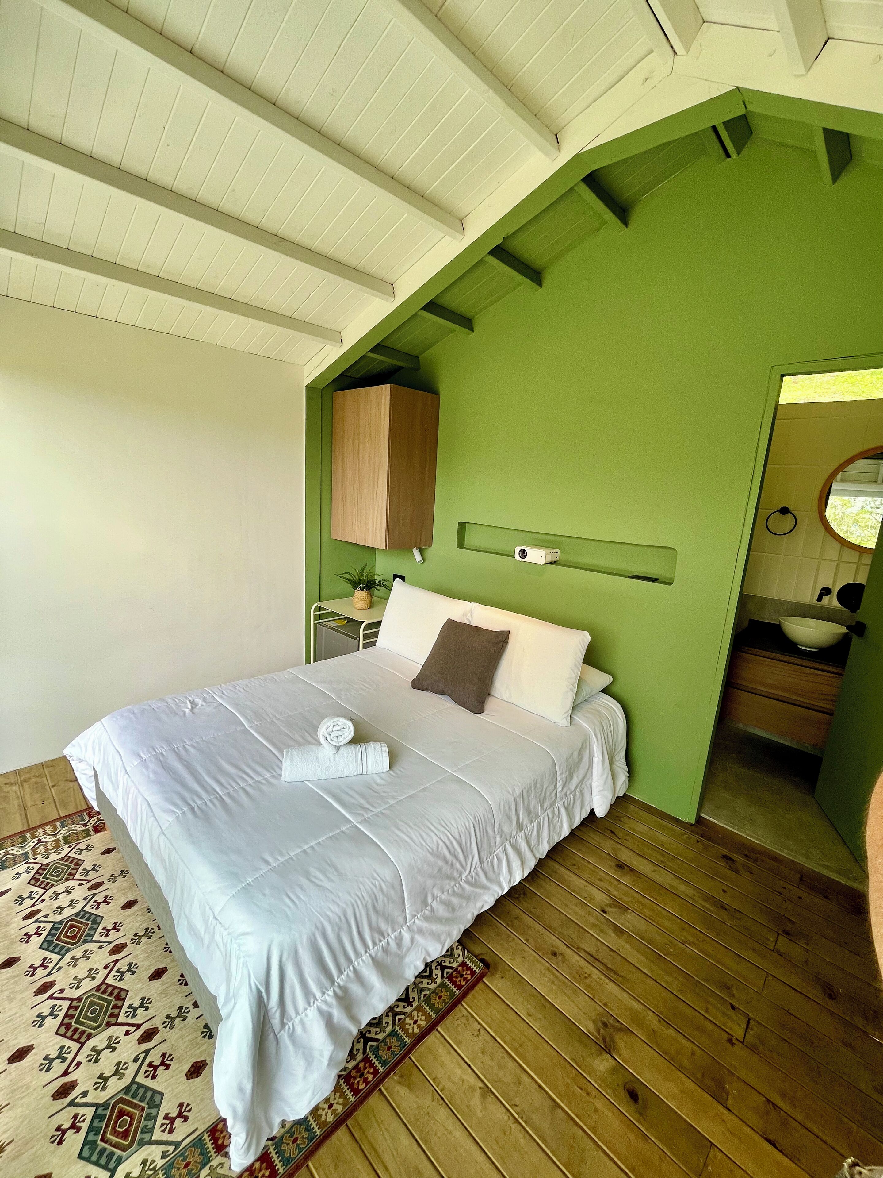 A bedroom with a bed, a green wall, a wooden floor, a small table with a plant, and a bathroom visible through an archway.