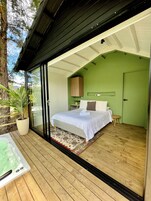 A modern bedroom with a large bed, a small bedside table, a potted plant, and a view of the outdoors.