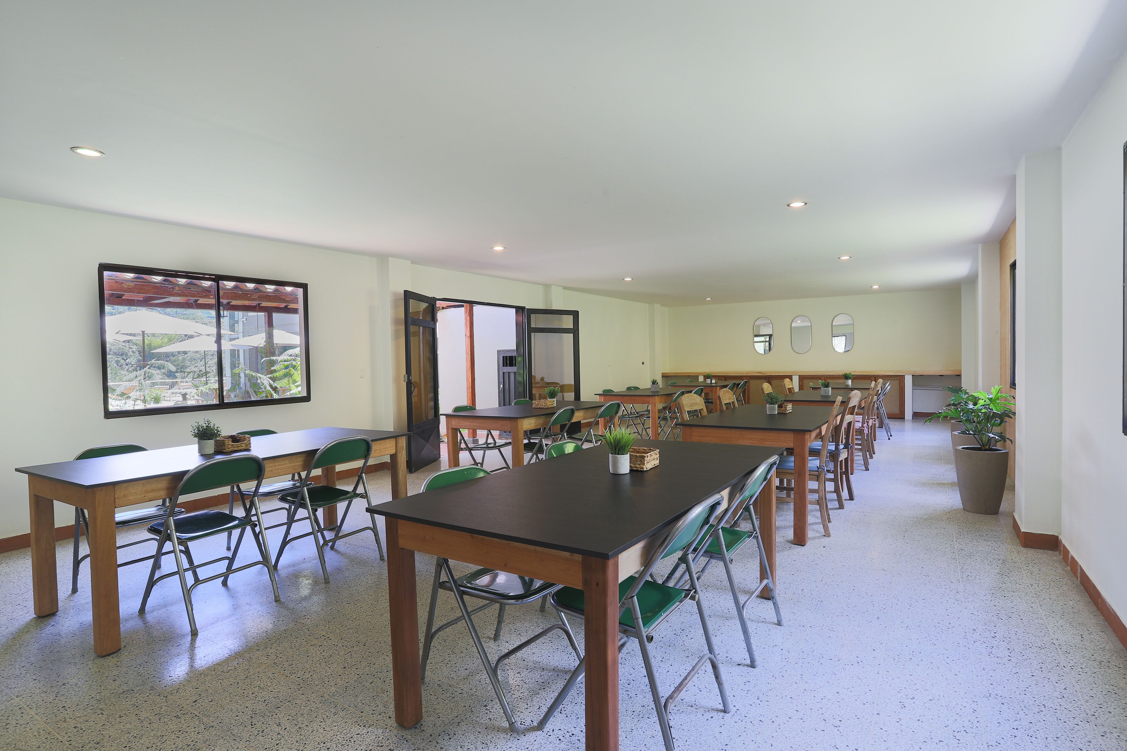 A spacious dining area with multiple tables and chairs, a large window with a view, and a potted plant.