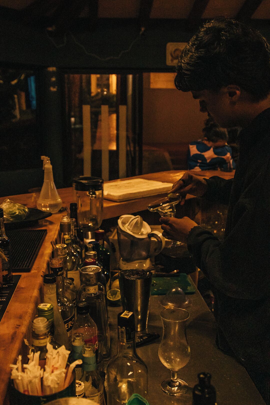 A bar with various bottles and a blender, a person preparing a drink, and a child in the background.