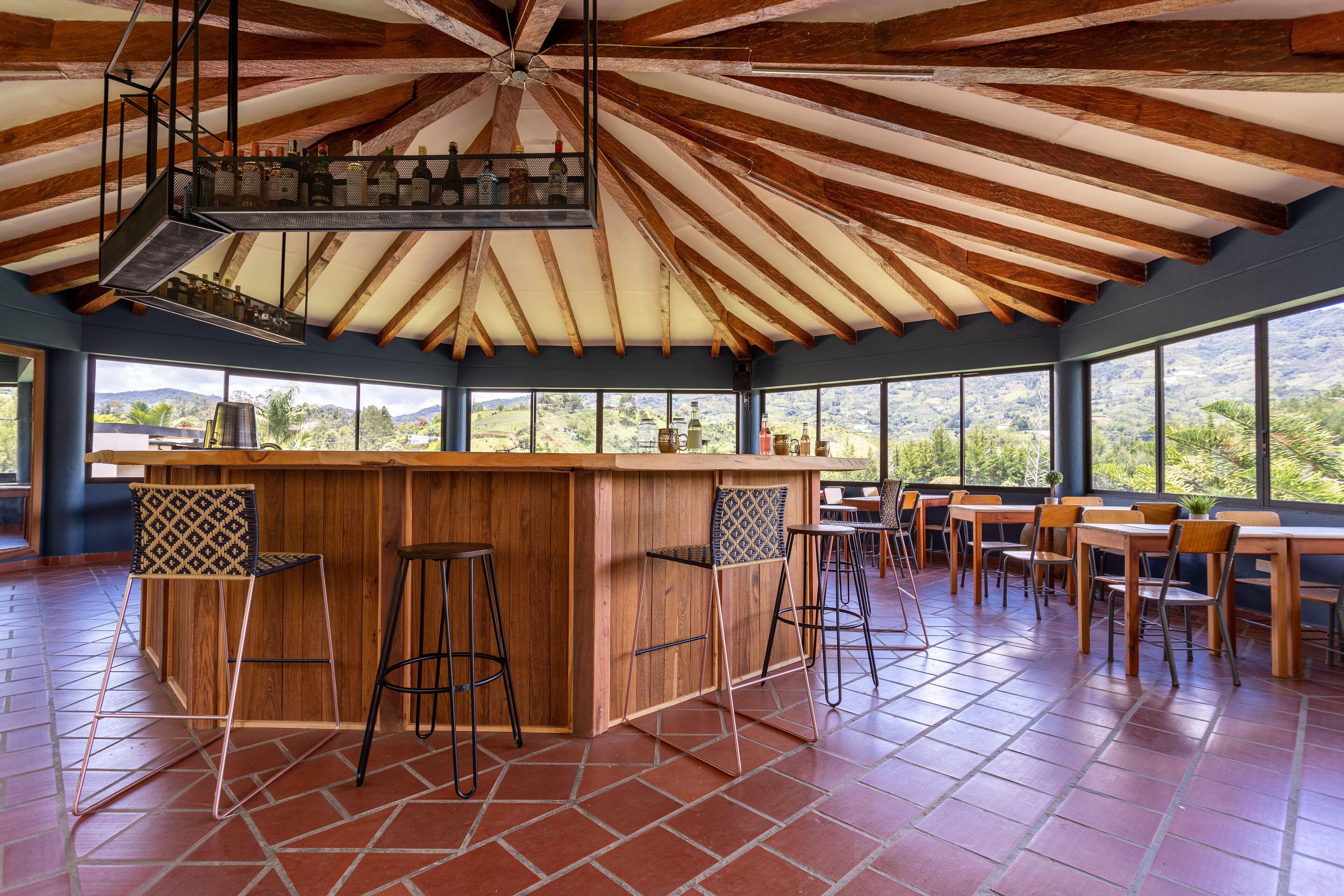 A bar area with wooden beams, a bar counter, stools, and large windows offering a view of the landscape.