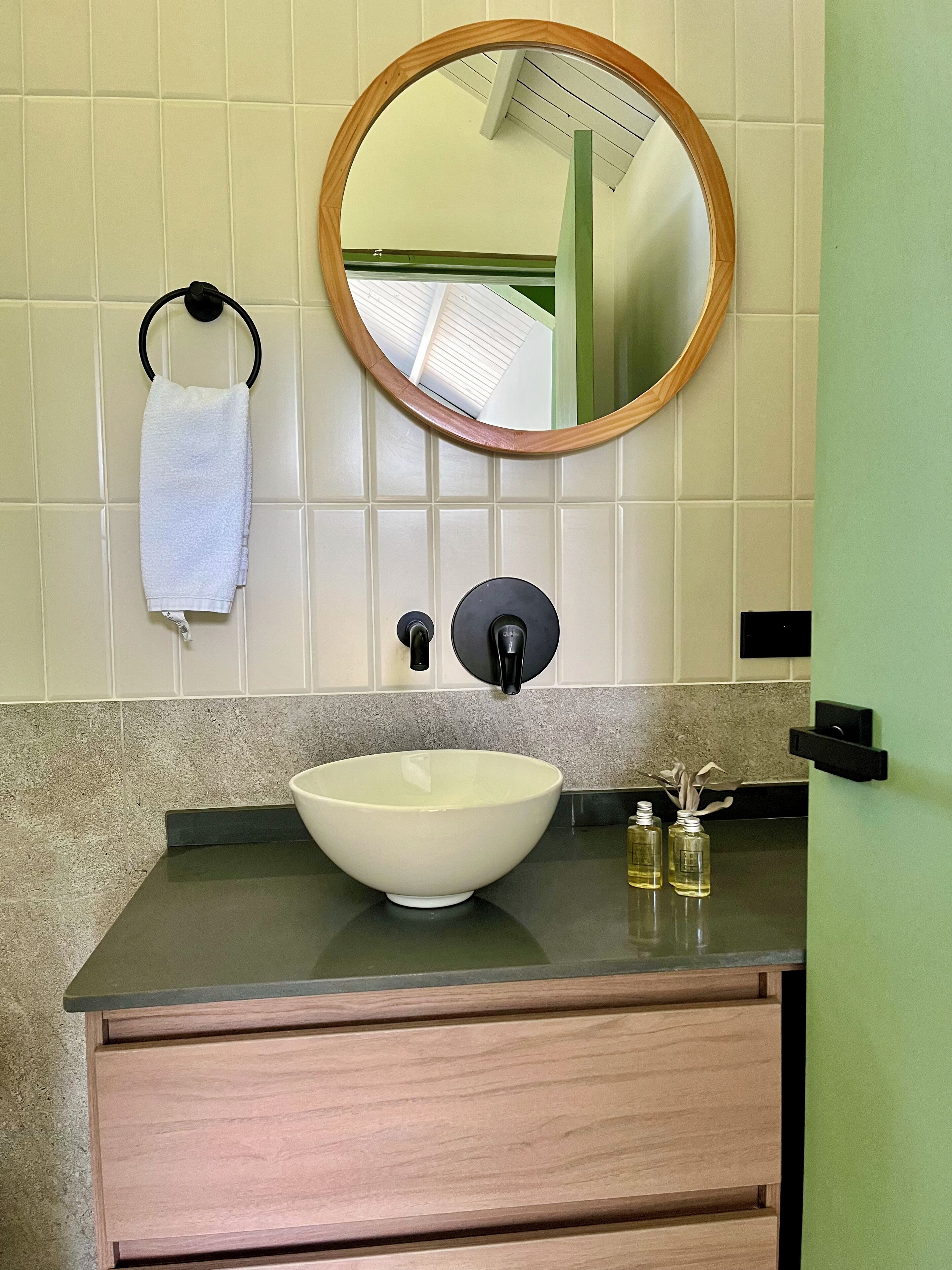 Casa de banho com lavatório branco, espelho redondo, porta-toalhas e porta verde.