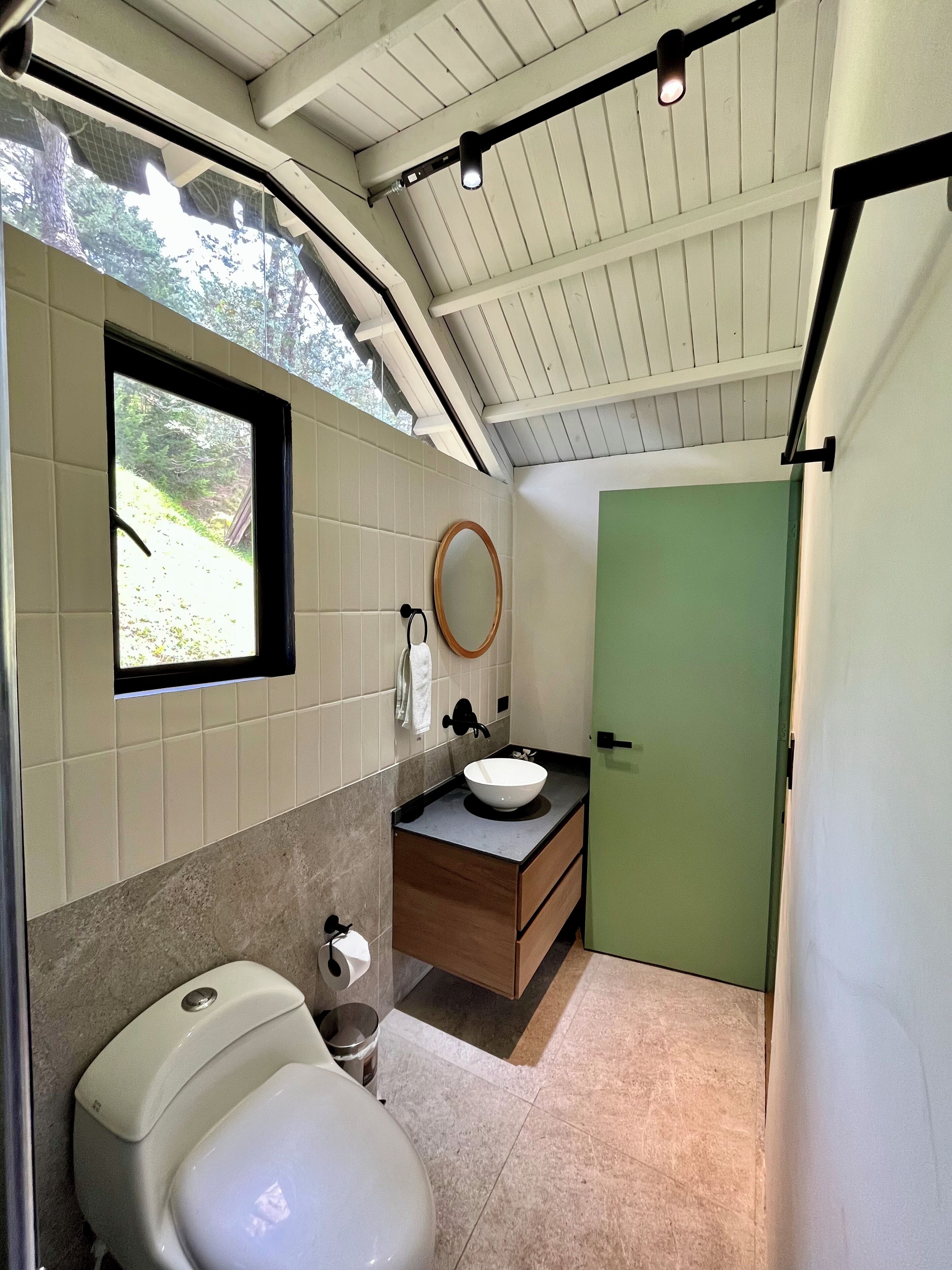 A modern bathroom with a toilet, sink, and mirror. There is a window providing natural light and a green door.