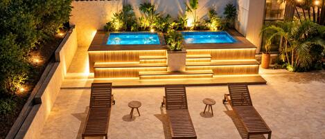 Ein Außenpoolbereich mit zwei Whirlpools, umgeben von Holzliegen und kleinen Tischen, mit Pflanzen und stimmungsvoller Beleuchtung.