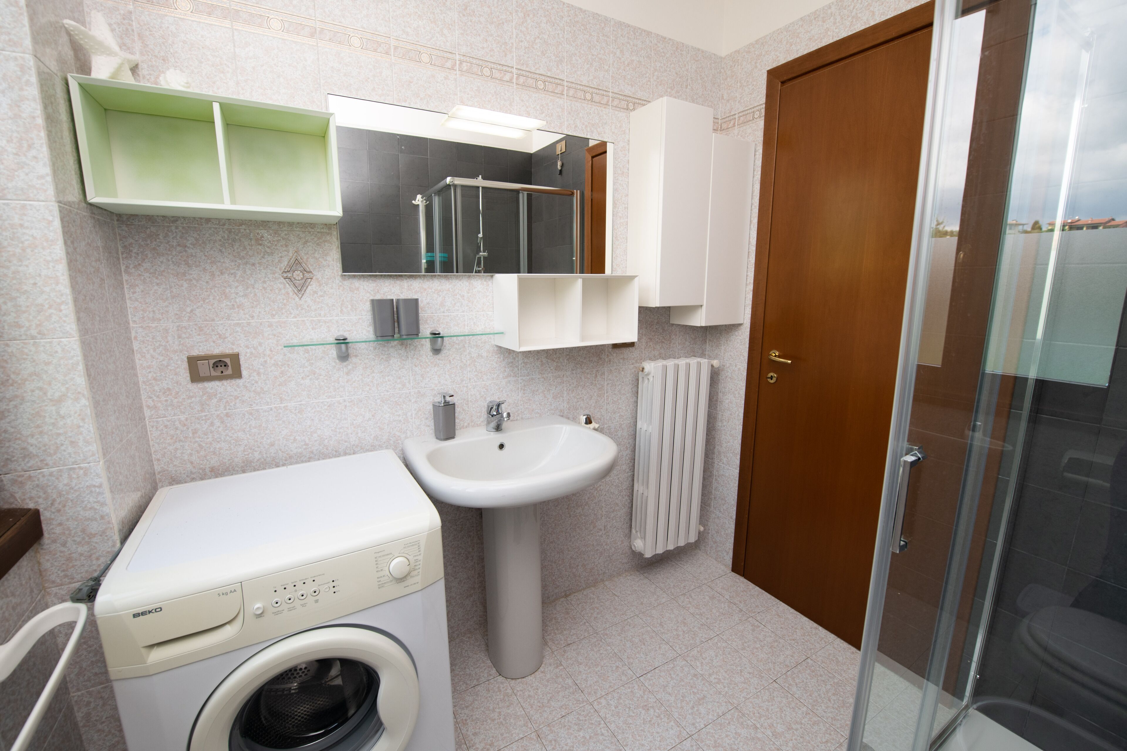 A compact bathroom with a washing machine, a sink, a mirror, and a glass-enclosed shower.