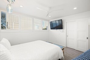 Ein Schlafzimmer mit einem Bett, einem an der Wand montierten Fernseher, einem Deckenventilator und einem Fenster mit Jalousien.