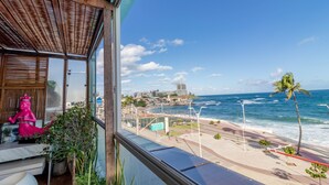 Une vue depuis un balcon donnant sur une plage, avec une statue, un palmier et, au loin, le paysage urbain.