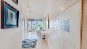 Un couloir moderne avec un sol carrelé blanc, un tapis à motifs bleus et un bureau blanc avec une chaise. Les murs sont ornés de tableaux encadrés et un grand miroir y est accroché.