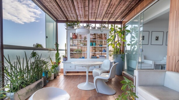 Un espace extérieur moderne aménagé avec une table ronde et des chaises, entouré de plantes en pot et doté d'une balustrade en verre donnant sur une étendue d'eau.