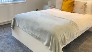 A bedroom with a large bed, a bedside table, a nightstand, a window with blinds, and a wall-mounted light. - Airport Comfy Stay (West Drayton)