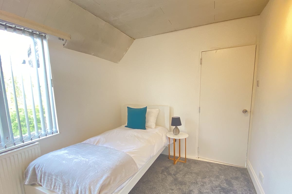 A small, single-bed room with a window, a bedside table with a lamp, and a door.