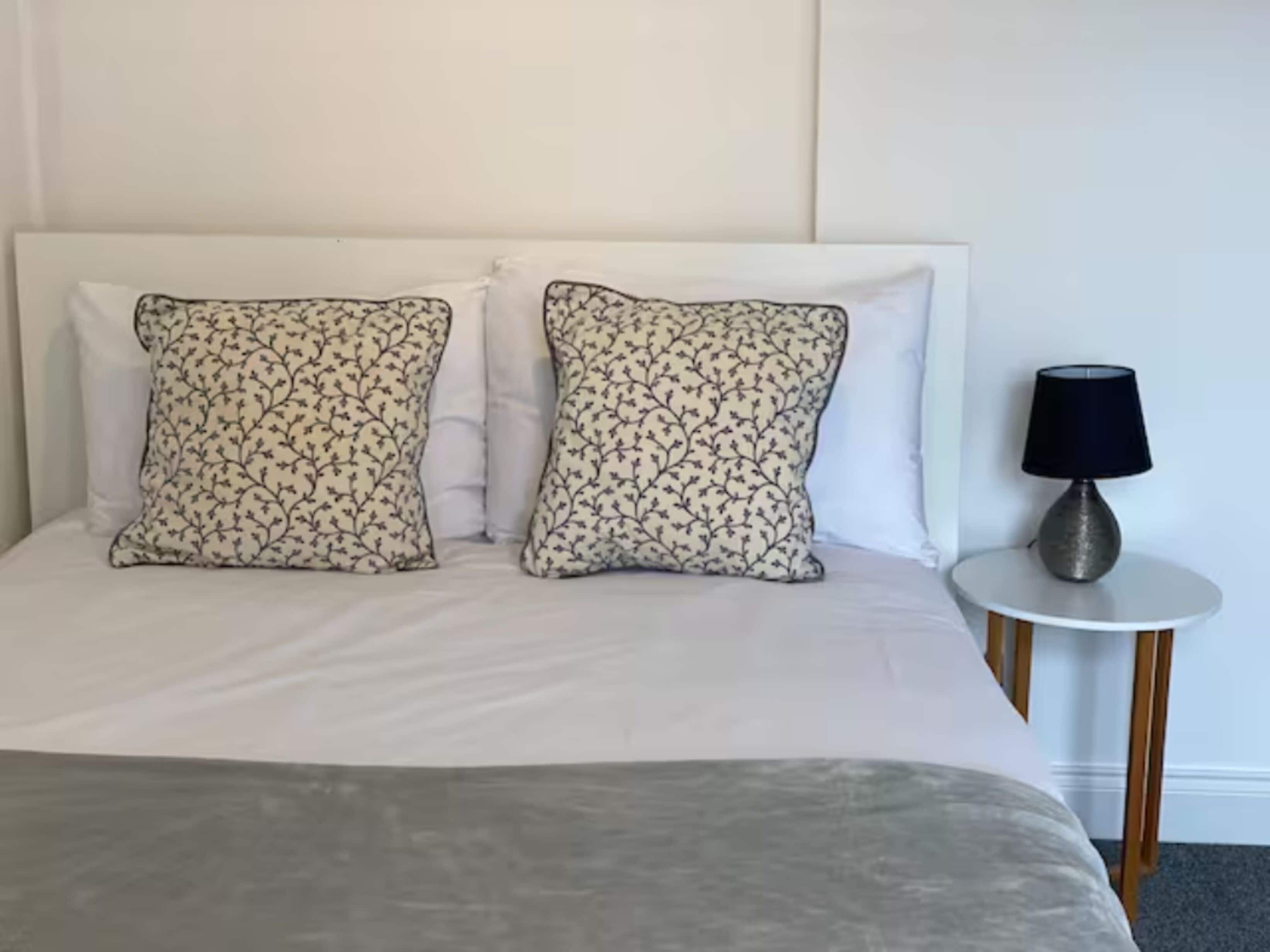 Un lit avec du linge de lit blanc, deux coussins à motifs et une petite table ronde avec une lampe à côté.