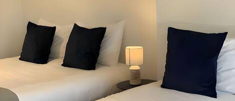 A hotel room with two beds, each with white bedding and black pillows, and a small table with a lamp between them.