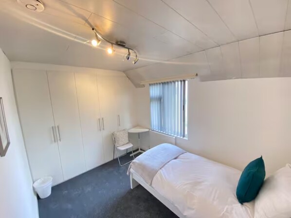 A bedroom with a bed, a window with blinds, a small table, and a wardrobe.