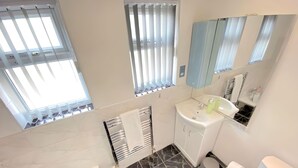 A bathroom with a white sink, mirror, and toilet. There are two windows with blinds, a towel rack, and a shower area. - Airport Comfy Stay (West Drayton)