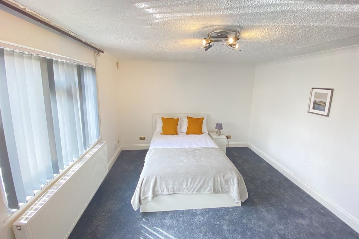 A hotel room with a bed, a nightstand, a lamp, a framed picture on the wall, and a window with blinds.