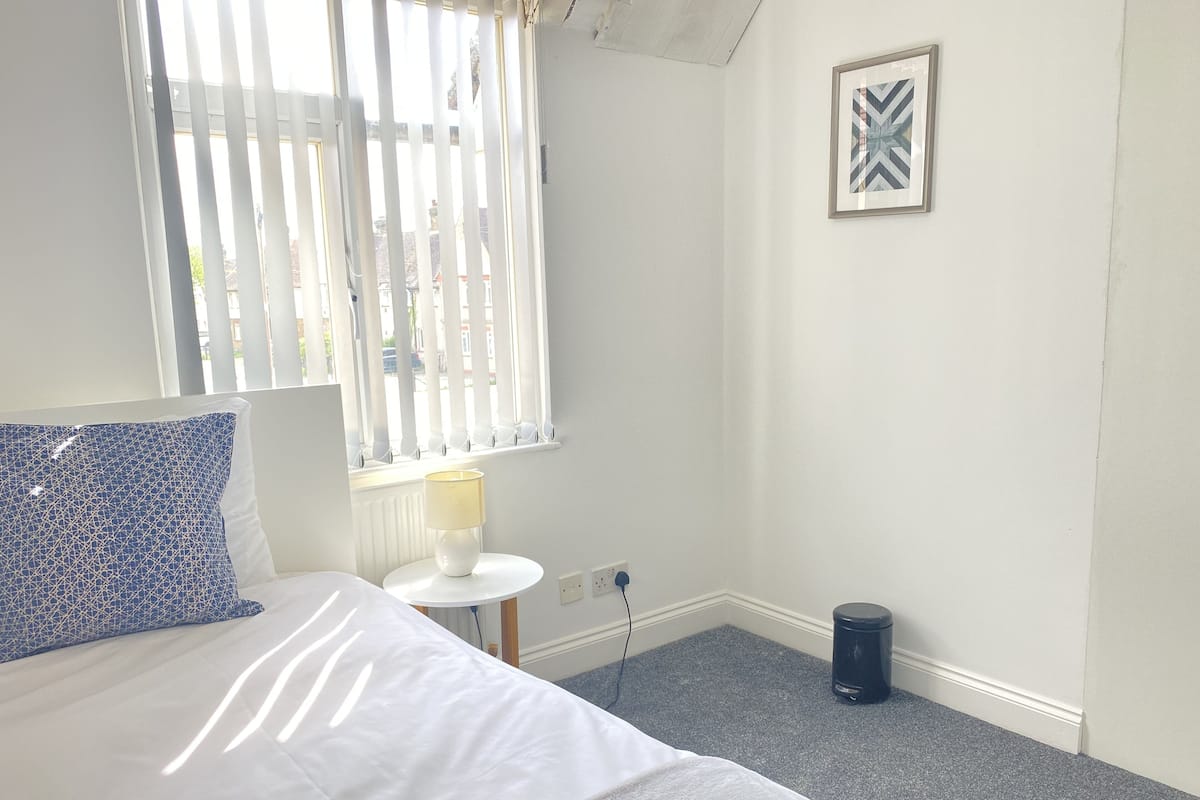 A bedroom with a bed, a small table with a lamp, a trash bin, and a window with blinds.