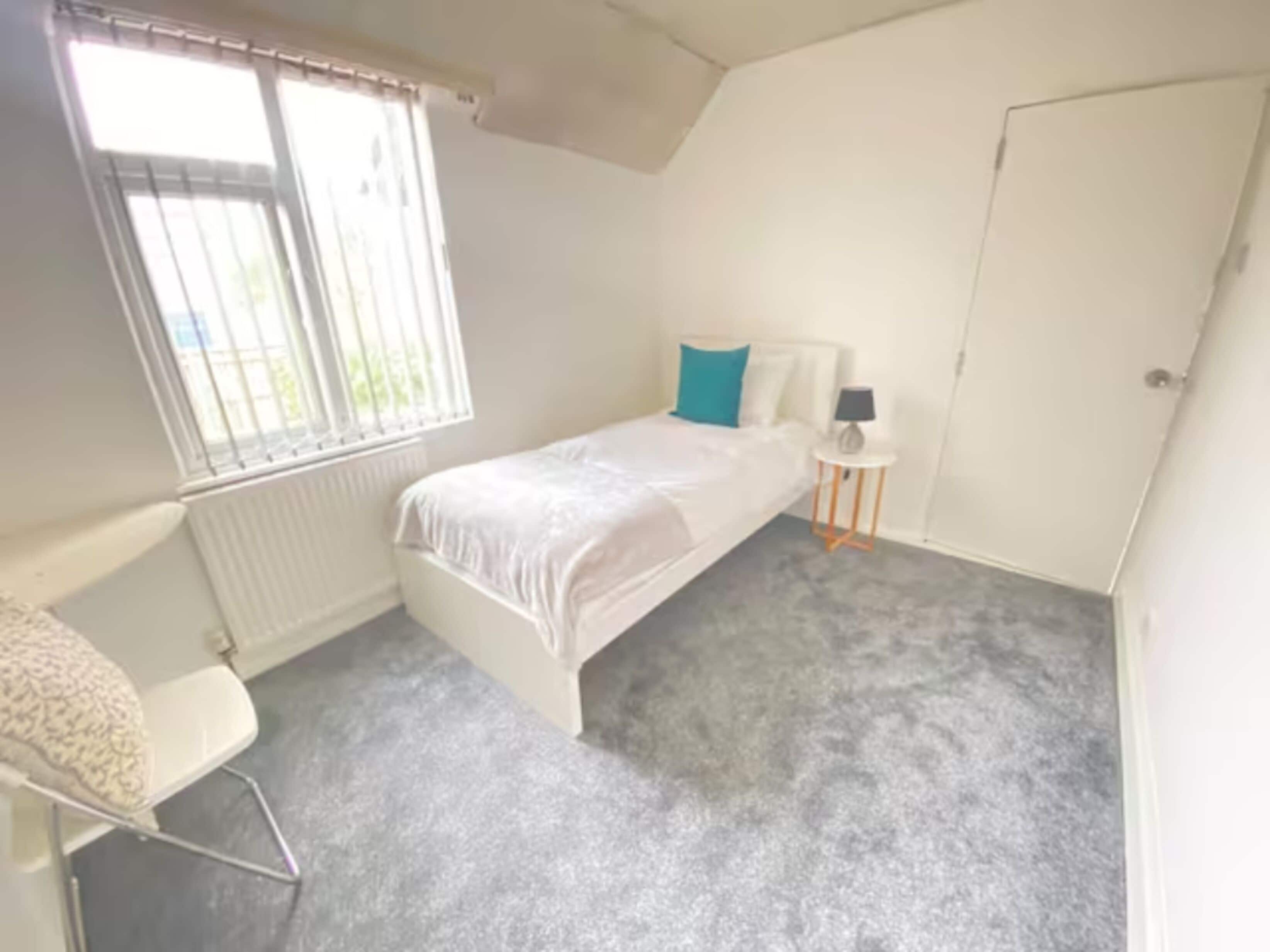 Un dormitorio pequeño y ordenado con una cama individual, una mesita de noche con lámpara y una ventana con persianas.