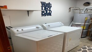 Une buanderie équipée de deux machines à laver et deux sèche-linge à chargement frontal, tous deux de couleur blanche, d’un chauffe-eau et d’une pancarte au mur indiquant : « Laver, sécher, plier, recommencer ».