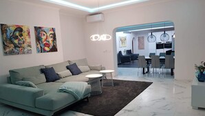 Un salon moderne avec un canapé d’angle, une table basse et un coin repas comprenant des chaises et une table. Deux tableaux ornent le mur et un luminaire est fixé au plafond.
