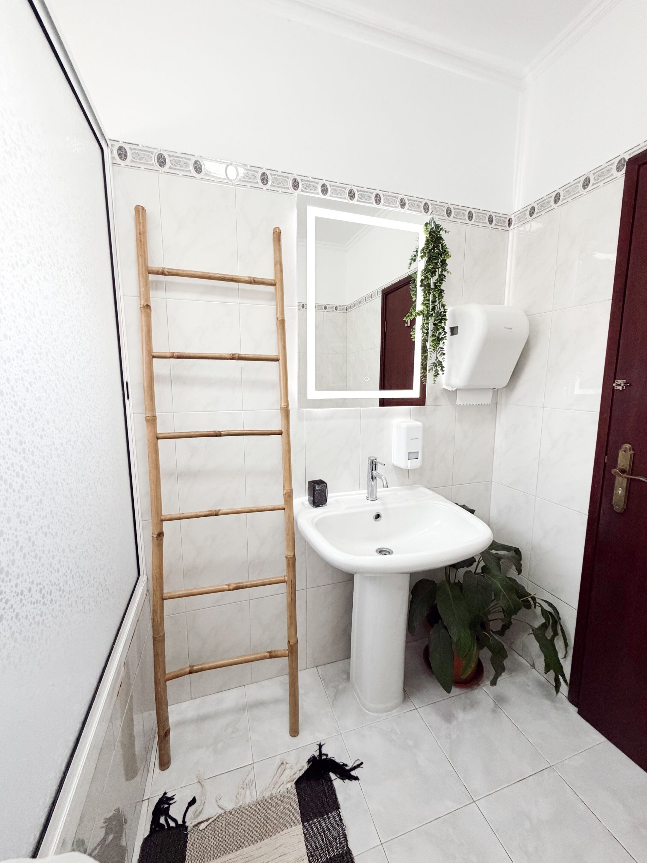 Une salle de bain avec un lavabo blanc, un miroir, une échelle en bois, une plante en pot et un sol carrelé.
