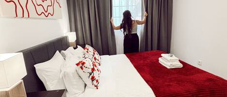 A modern hotel room with a bed, a bedside table, a red blanket, and a large window with curtains.