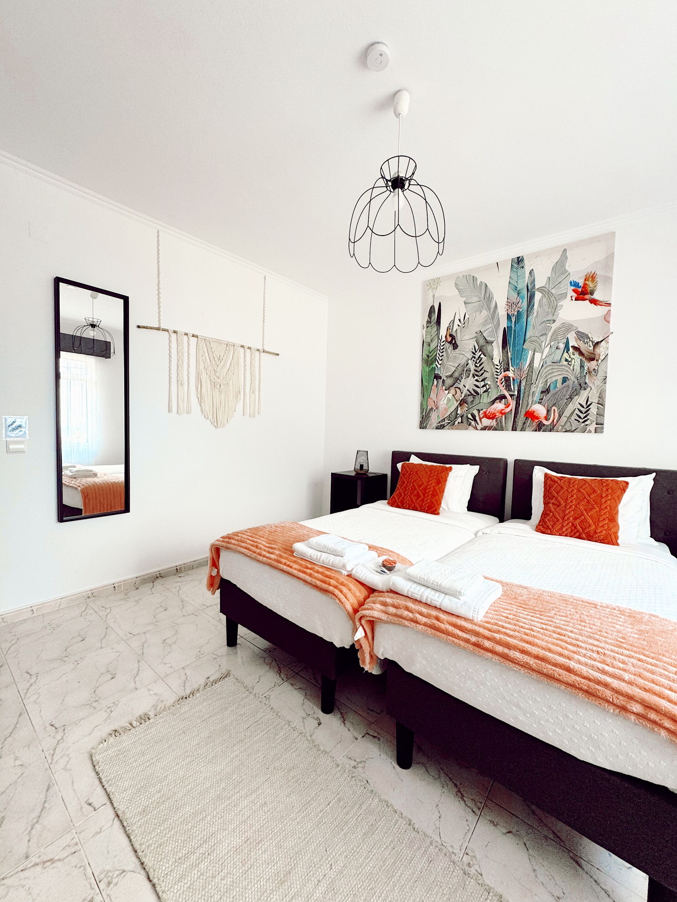 A modern bedroom with a large bed, a mirror, a macrame wall hanging, and a tropical-themed painting.