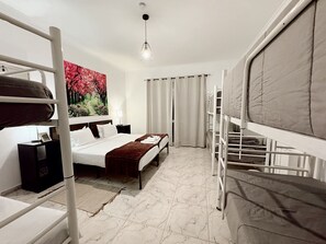 Ein Hotelzimmer mit Etagenbett, einem Einzelbett, einem Nachttisch mit Lampe, einem Bild an der Wand und einer Deckenleuchte.