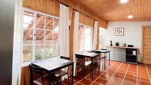 Ein geräumiges Zimmer mit Holzbalkendecke und Fliesenboden, ausgestattet mit einer Essecke mit Tischen und Stühlen, einer Küchenzeile mit Spüle und Schränken, sowie großen Fenstern für viel Tageslicht.