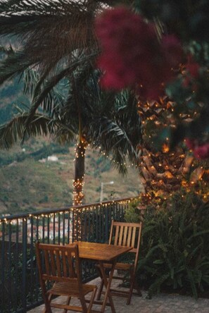 Un patio con mesa y sillas de madera, rodeado de follaje y palmeras, con luces colgantes.
