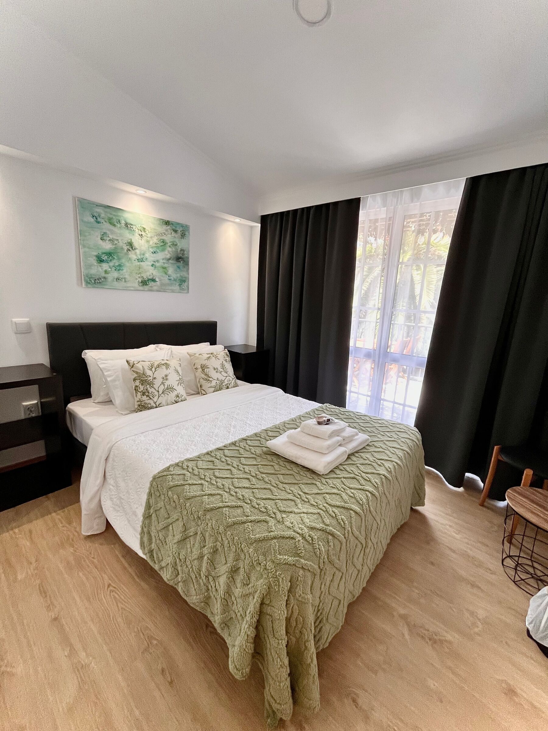 Ein modernes Schlafzimmer mit einem großen Bett, einem Nachttisch, einem Wandbild und Blick ins Freie durch ein Fenster mit Vorhängen.