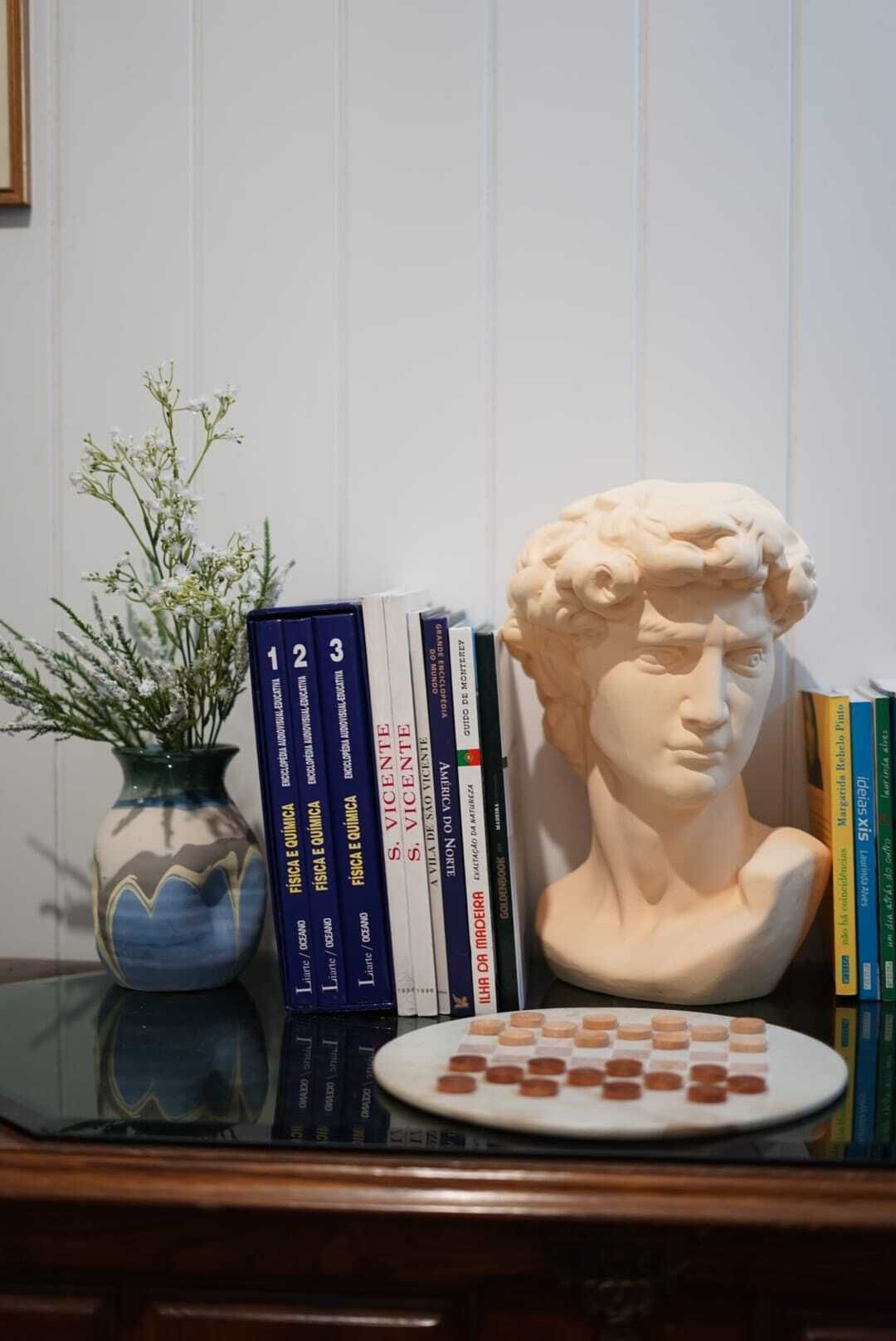 A bookshelf with books, a vase with flowers, a bust sculpture, and a plate with pills.