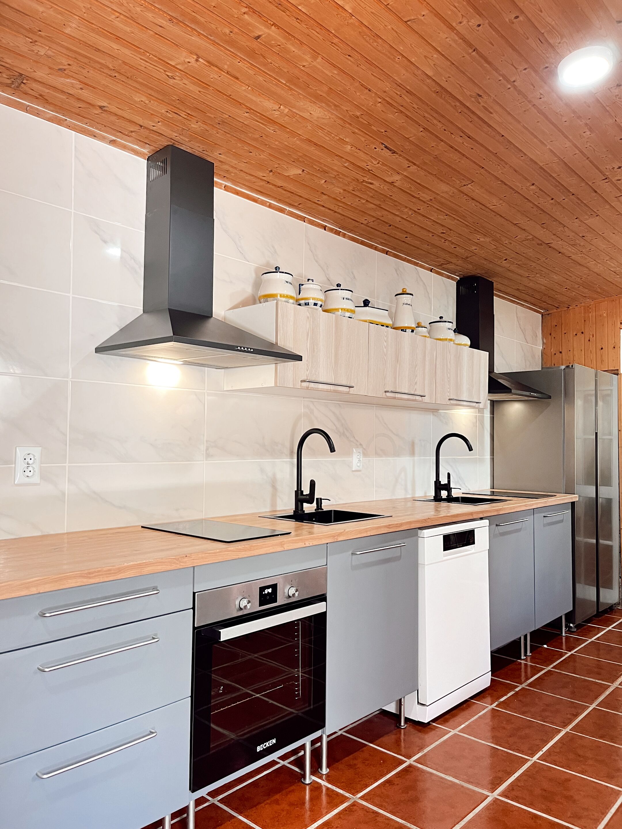 A modern kitchen with a wooden ceiling, white marble tiles, a stainless steel range hood, a double sink, a built-in oven, and a dishwasher.