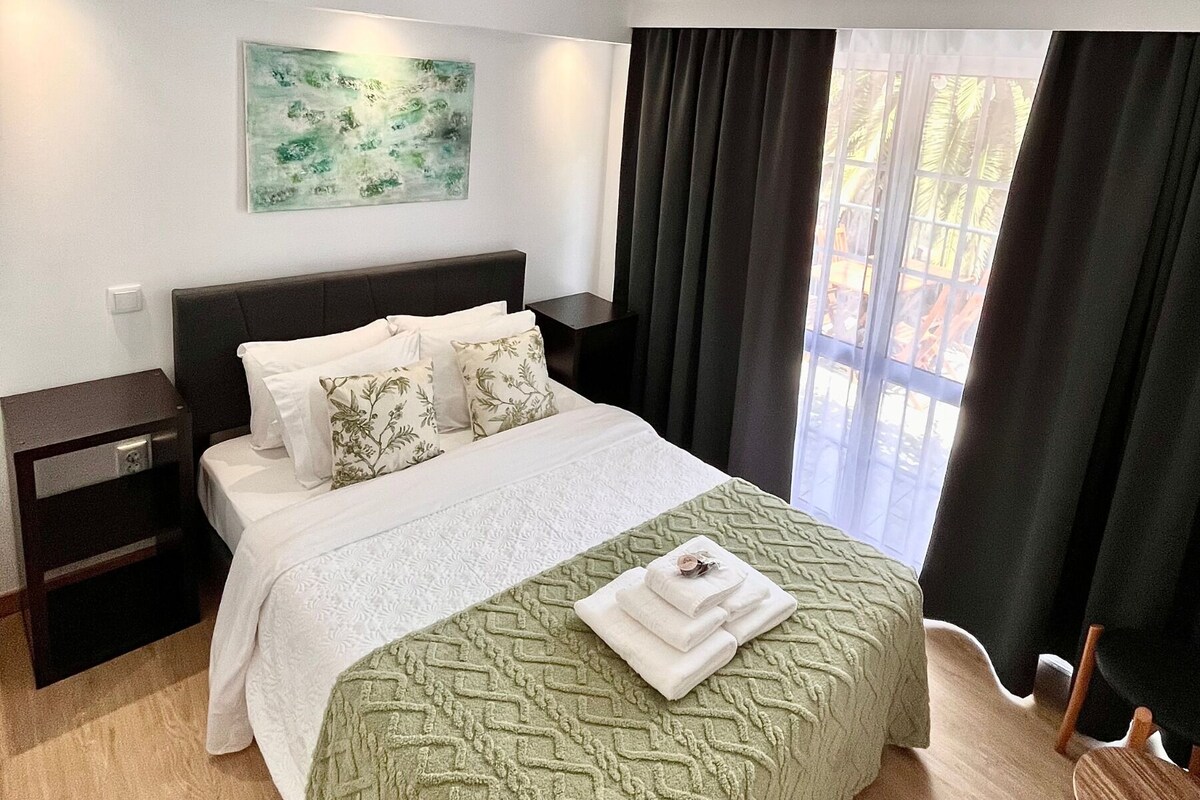 A modern bedroom with a large bed, a nightstand, a wall-mounted artwork, and a view through a window with curtains.