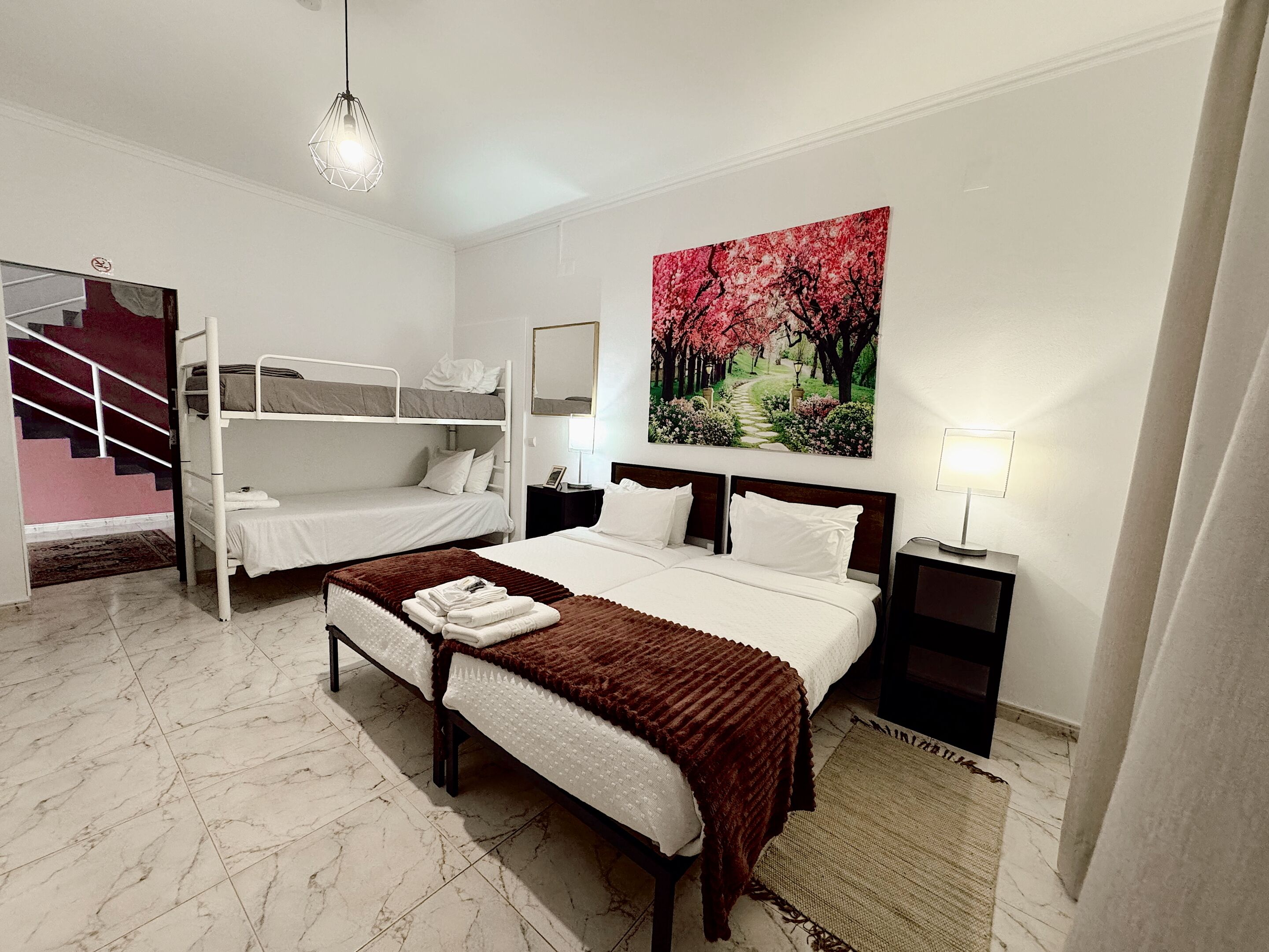 Habitación de hotel con literas, una cama individual, un cuadro de un sendero de cerezos, una lámpara de noche y un espejo en la pared.