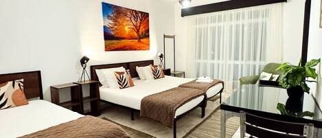 A hotel room with two beds, a dining table, a potted plant, and a large window with sheer curtains.