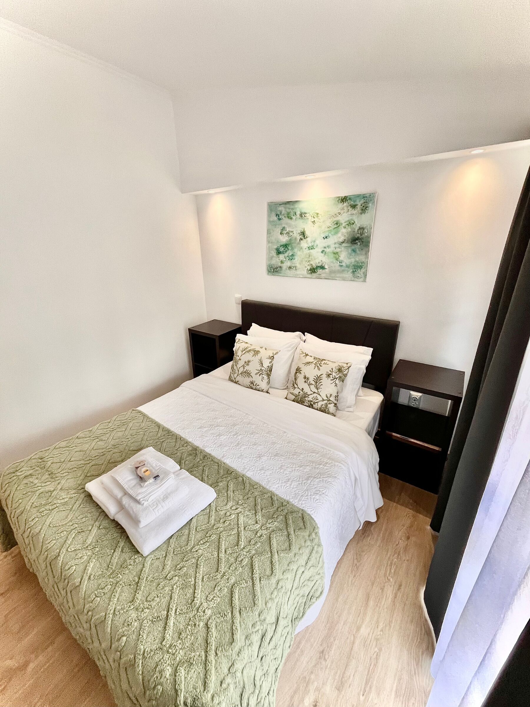 A neatly made bed with a green quilt, white pillows, and a folded towel on the bed. There is a nightstand with a lamp and a small drawer. A painting hangs on the wall above the bed.