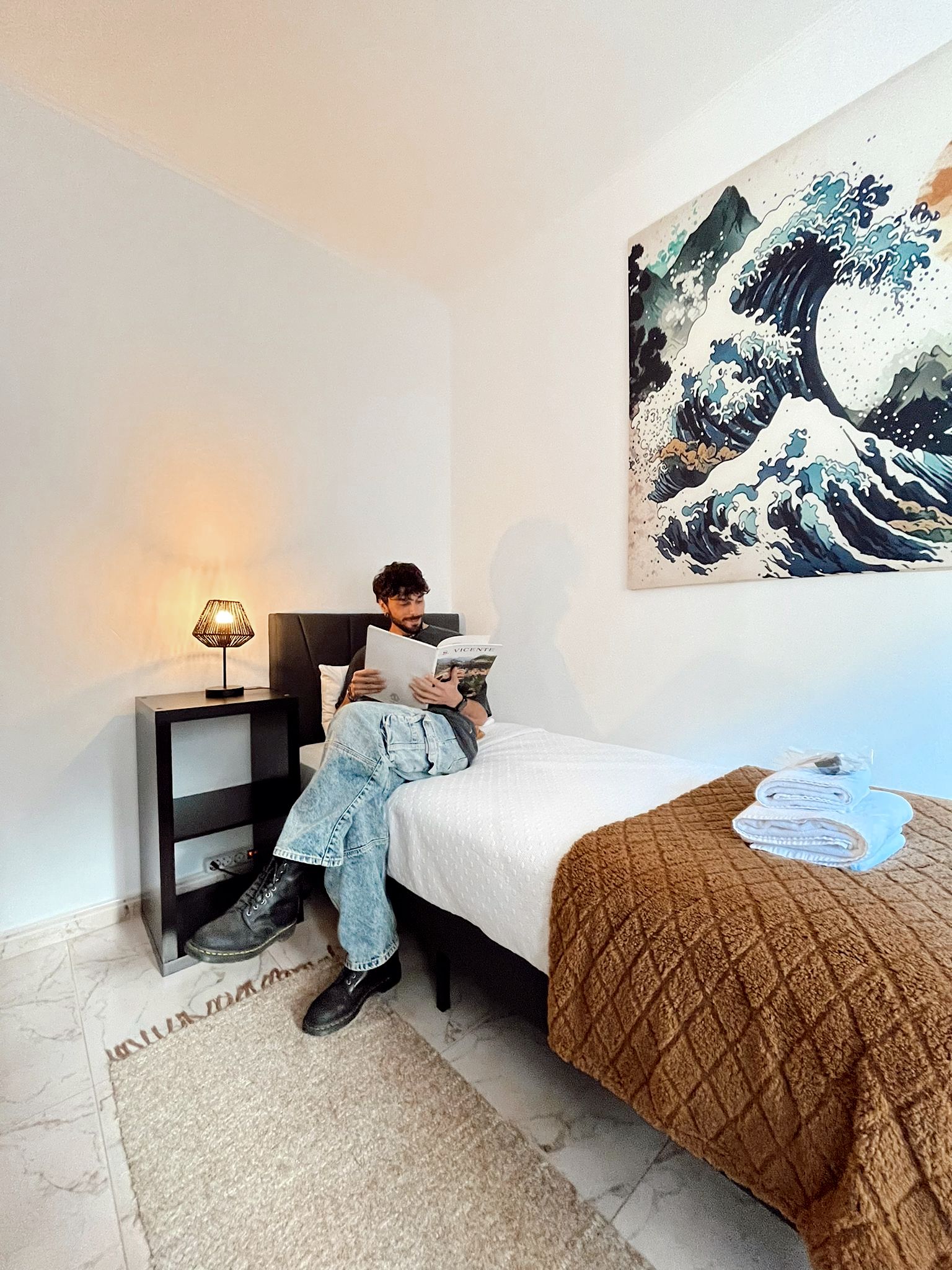 A bedroom with a bed, a nightstand, a lamp, a blanket, and a painting on the wall.